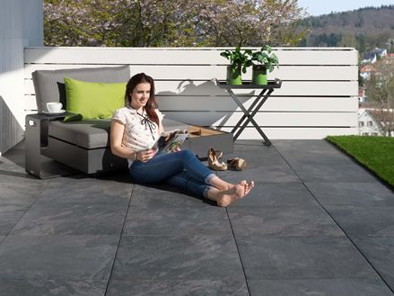 Junge Frau liest auf Terrasse mit dunklen Feinsteinzeugplatten in Schieferoptik und modernen Möbeln
